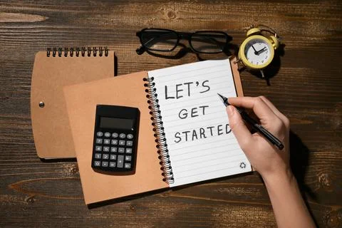 Female hand writing text LET'S GET STARTED in notebook on wooden background Stock Photos