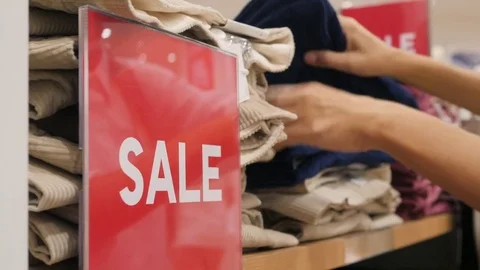 Female Hands Choosing Pants from Stack in Clothing Store. Big Red Sign in Stock Footage 82106090
