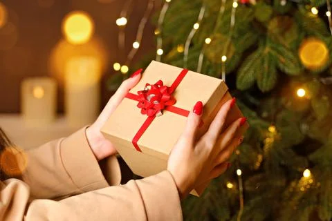 Female hands holding craft gift box with red ribbon near Christmas tree Stock Photos