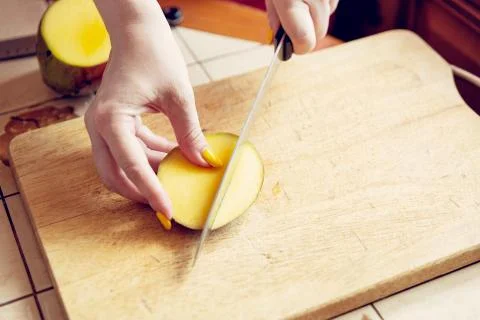 Female hands with a knife cut a mango on a cutting board. Stock Photos