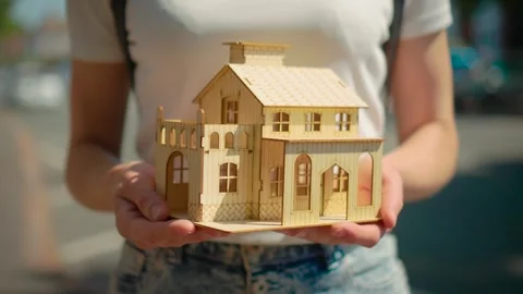 Female hands presenting tiny cottage model on city street. Housing loan and Stock Footage 336969503