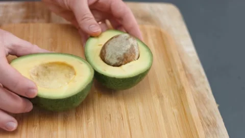 Female hands separating two halves of avocado on a wooden cutting board. Cooking Stock Footage 150500816
