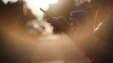 Female hands using smartphone and holding paper cup of coffee on sunset Видео 108410294