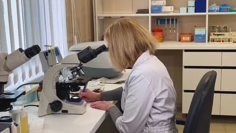 Female laboratory assistant looking through microscope and writing down data on Stock Footage 270279976