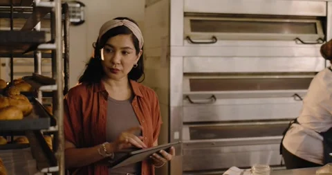 Female manager and chef in white coat standing by oven bank, tapping tablet Stock Footage 337026569