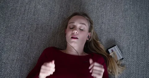 Female maroon top lying on gray carpet listening with wired buds to phone, Stock Footage 344692860