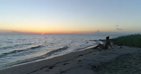 Female model sitting on a beach watching a beautiful sunset over the water, aeri Stock Footage 134911222