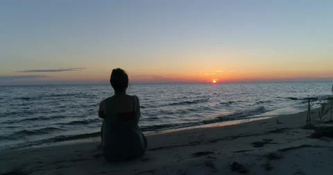 Female model sitting on a beach watching a beautiful sunset over the water, aeri Video stock 134990071