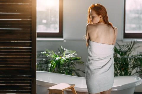 Female model standing between window and bathtub, using towel to hide her nudity Stock Photos