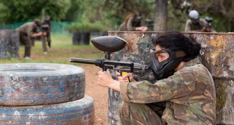 Female paintball player in camouflage and mask aiming with gun in shootout Stock Photos