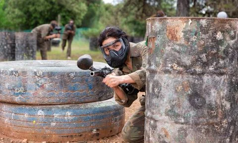 Female paintball player in camouflage and mask aiming with gun in shootout Фото