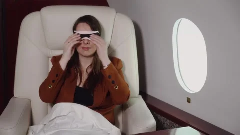 Female passenger in brown jacket adjusts sleep mask while seated in luxurious Video stock 328480892
