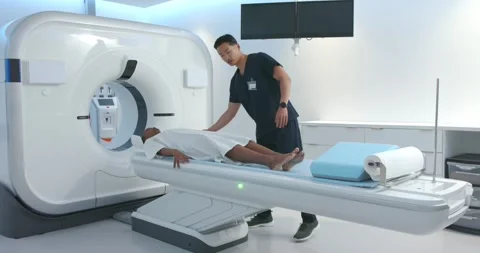 Female patient lying on scanner table, Asian tech placing blue wedge under Stock Footage 336483997
