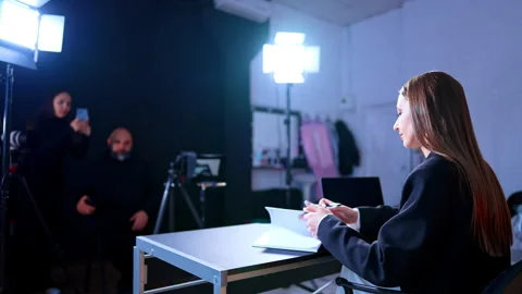 Female presenter in black blazer at desk during video production. Woman sits at Video stock 329192537