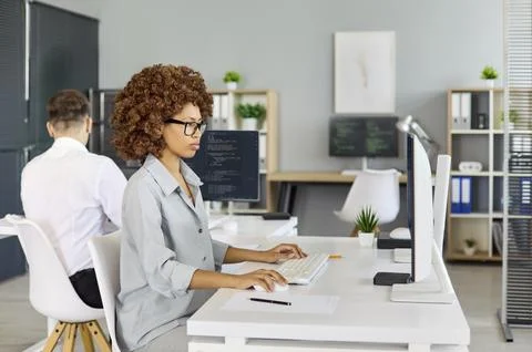 Female programmer coding at desktop in modern office with coworker in background Stock Photos