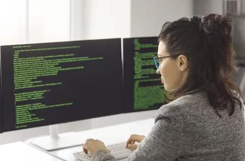 Female programmer coding on dual computer screens in modern office workspace Stock Photos