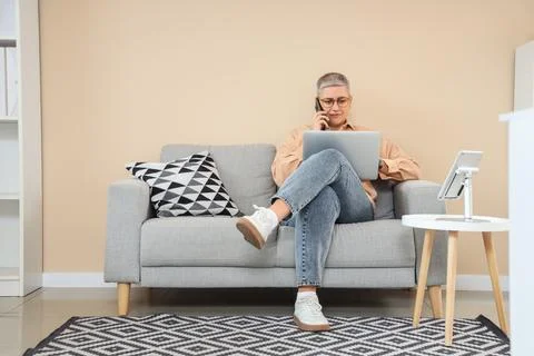 Female programmer with laptop talking by mobile phone on sofa in office Stock Photos