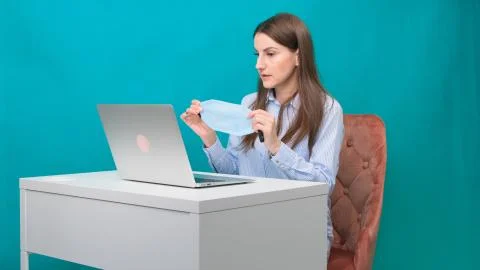 Female puts on a protective mask while working on a laptop in the workplace or Stock Photos