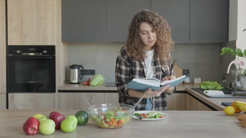Female Reading A Book While Standing In Kitchen With Healthy Salad Nearby Stock Footage 311282703