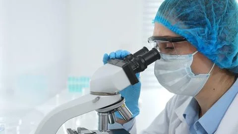 Female scientist analyzing sample through microscope, wearing protective lab Stock Photos