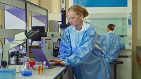 Female scientist using a laptop while a colleague works in a cabinet Vídeo Stock 346940008