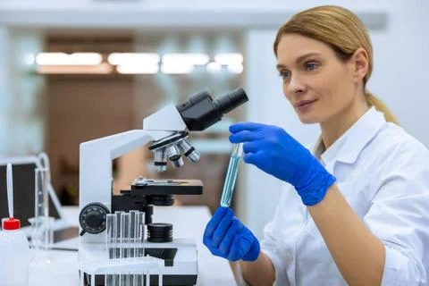 Female scientist using microscope, holding test tube, analyzing liquid bio... Stock Photos