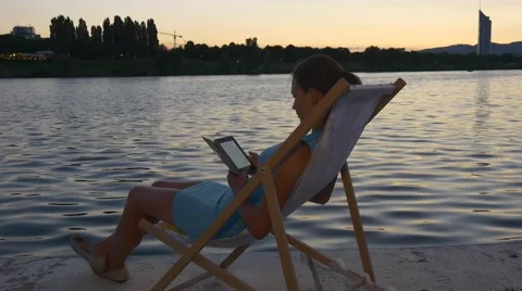 Female sits on a sunbed reading e-reader at the waterfront Stock Footage 66431032