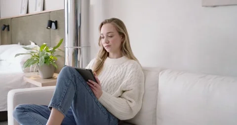 Female sitting on light sofa in small living room, holding tablet, producing Stock Footage 344680679