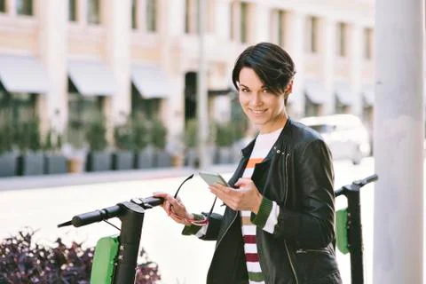 Female stands by electric scooter in the city and looks at camera smiling - C Stock Photos