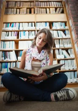 Female student against bookshelf reading a book on the library floor Stock Photos