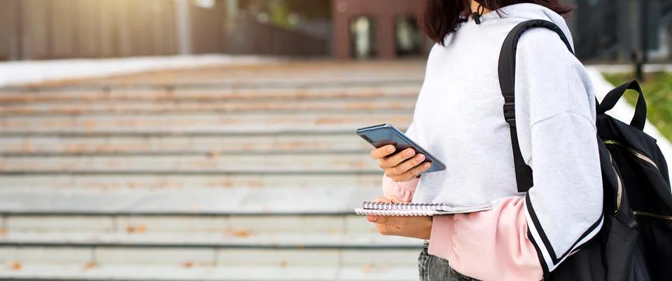A female student with a black backpack holds a smartphone and Notepad on the  Stock Photos