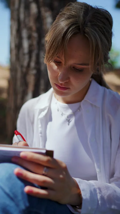 Female student scribbling notes with red pen, leaning against pine tree trunk Stock Footage 308382242