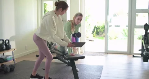 Female trainer adjusting tablet while guiding client rowing dumbbell on bench in Stock Footage 335454220