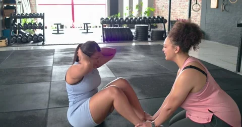 Female trainer-client doing sit-ups in gym on cue holding ankles counting for Stock Footage 344224318