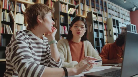 Female University Students Discussing Homework on Laptop in Library Stock Footage 277709547