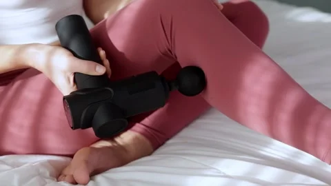Female using a vibrating massage gun on her thigh while seated on a bed, Stock Footage 325135852