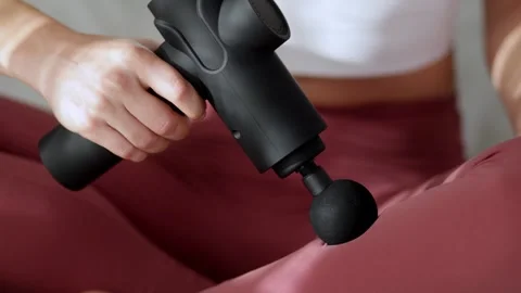 Female using a vibrating massage gun on her thigh while seated on a soft Stock Footage 325136143
