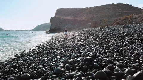 Female is walking by the ocean on a beach full of stones and wets her legs in Stock Footage 94124732