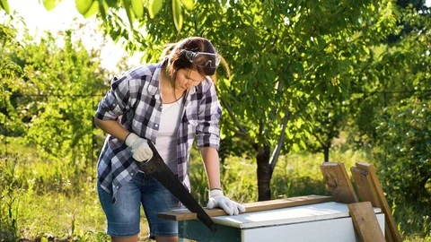 Female woodworker in checked shirt sawing wooden plank in summer garden Stock Footage 112946853