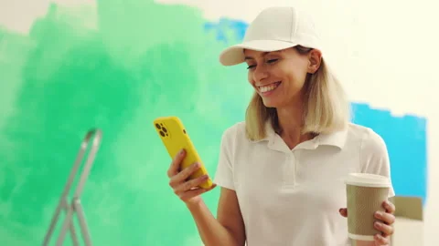 Female worker standing, using smartphone after painting the wall and drinking Stock Footage 229396746