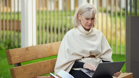 Female writer types on laptop while sitting on bench near greenery. Thoughtful Stock Photos