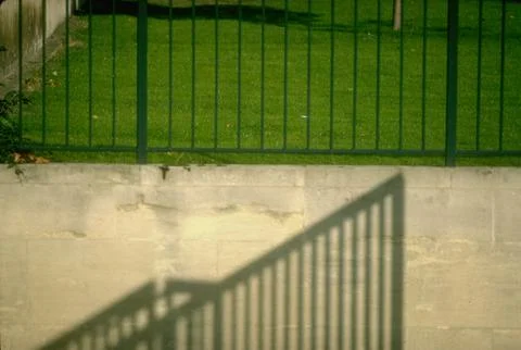 Fence framing grassy plot appearing to cast shadow on low stone wall beneath., L Fotos de archivo