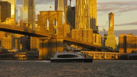 Ferry on East River crosses under Brooklyn Bridge at dawn, skyscrapers behind Stock Footage 99091663