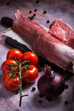 Few red cherry tomatoes next to pork tenderloin on wooden board Stock Photos