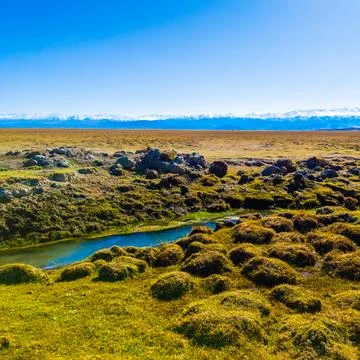 Field covered with dry grass bumps and small creek with distant high mountains Stock Photos