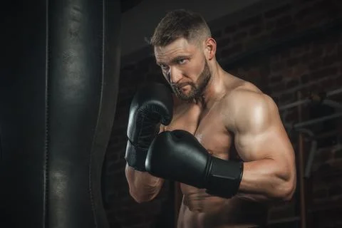 Fighter with aggression beats punching bag in black boxing gloves in the gym. 库存照片