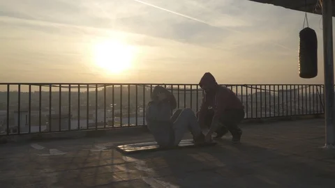 Fighter doing crunches on terrace, trainer holding his feet, boxing bag, sunset. Stock Footage 98479227