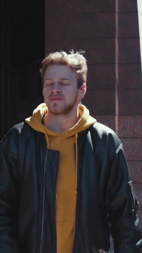 A figure in a leather jacket and yellow hoodie stands before a building Stock Footage 314172407