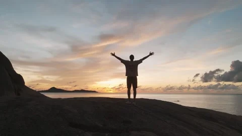 Figure on ocean coast at orange sunset raising arms in delight grandeur of Stock Footage 312505012