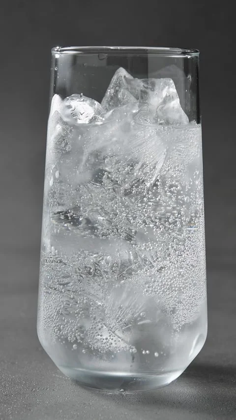 Fine bubbles rise through clear iced drink in glass on gray studio background, Stock Footage 345486745